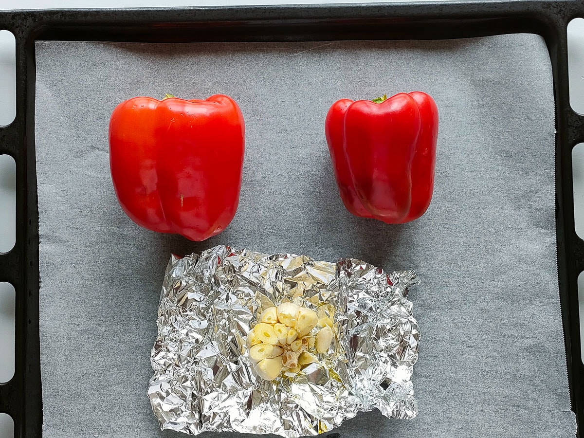 2 red peppers and garlic drizzled with oil on a sheet pan lined with parchment paper.