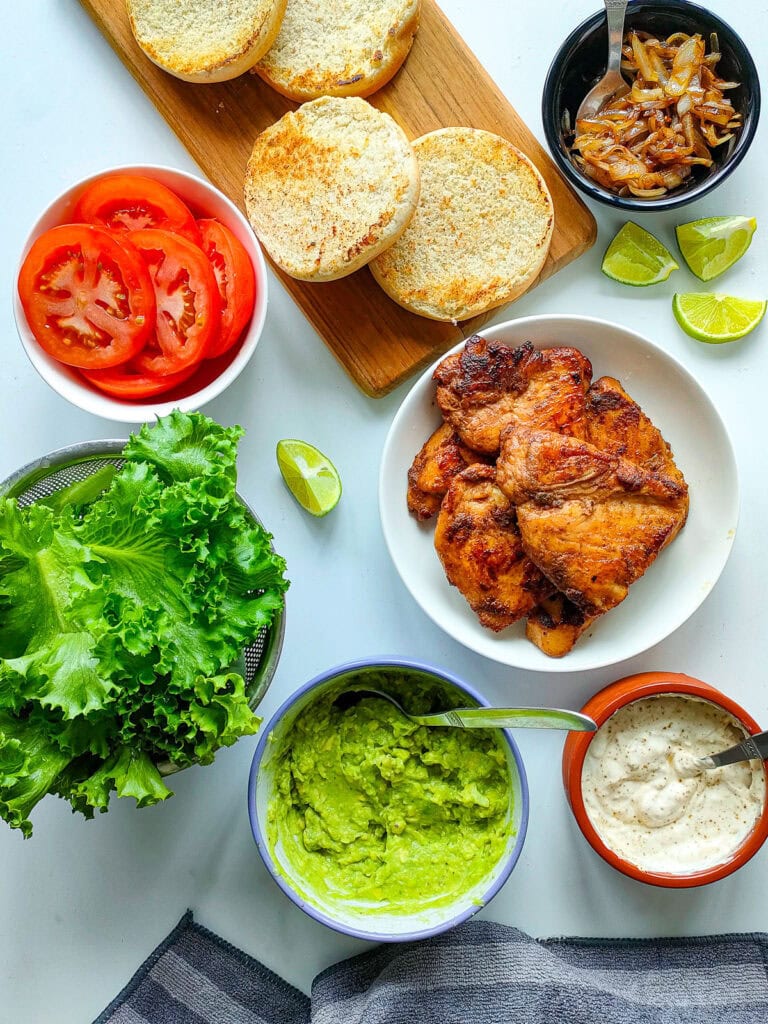 Ingredients for chipotle chicken sandwich: chipotle lime chicken, yogurt sauce, avocado spread, veggies and toasted buns in separate bowls.