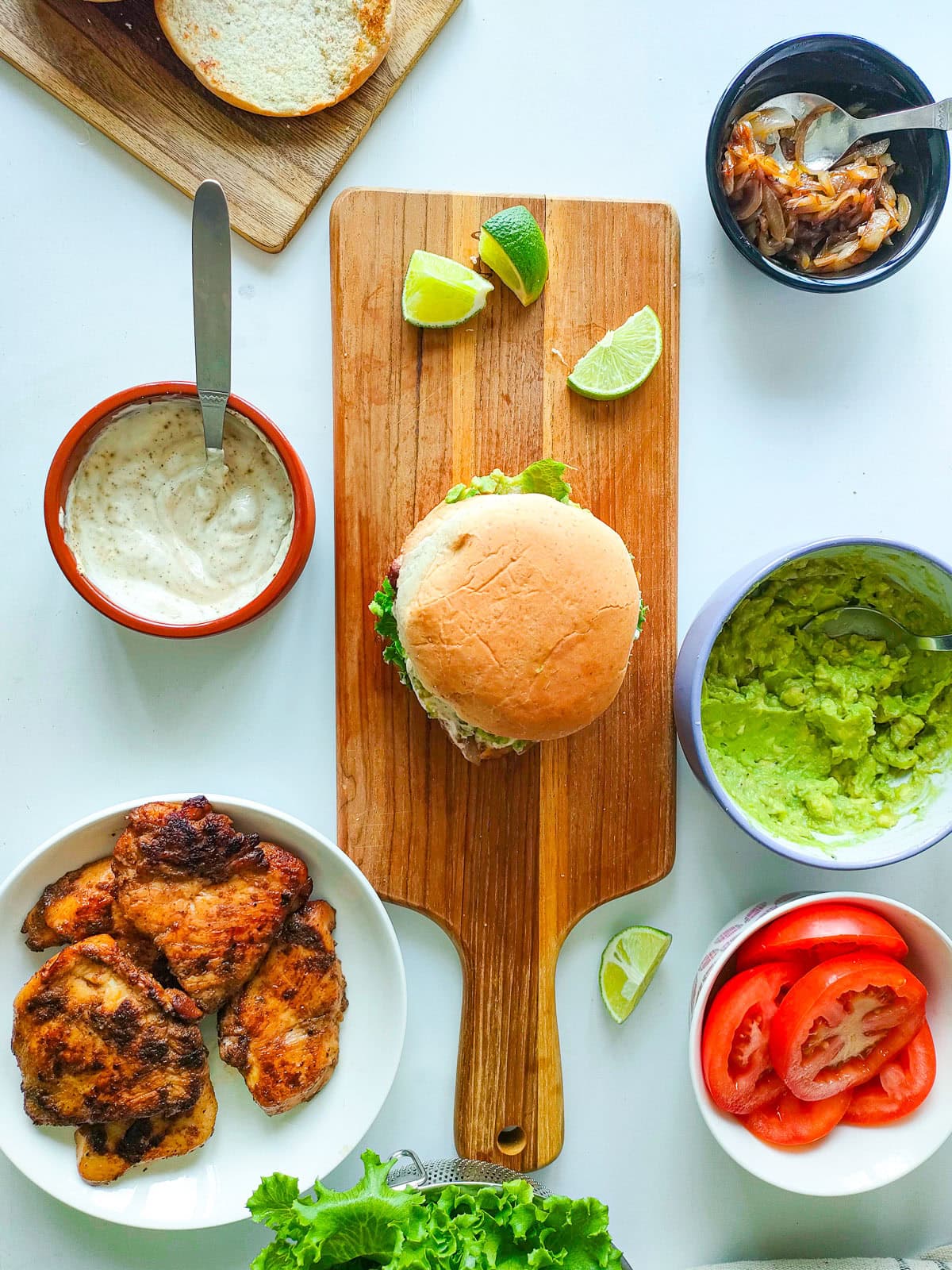 Wholesome Chipotle Chicken Sandwich with Easy Avocado Spread Chipotle chicken sandwich on a serving board with lime wedges and a plate of chipotle lime chicken and spread, sauce and veggies in separate bowls next to it.