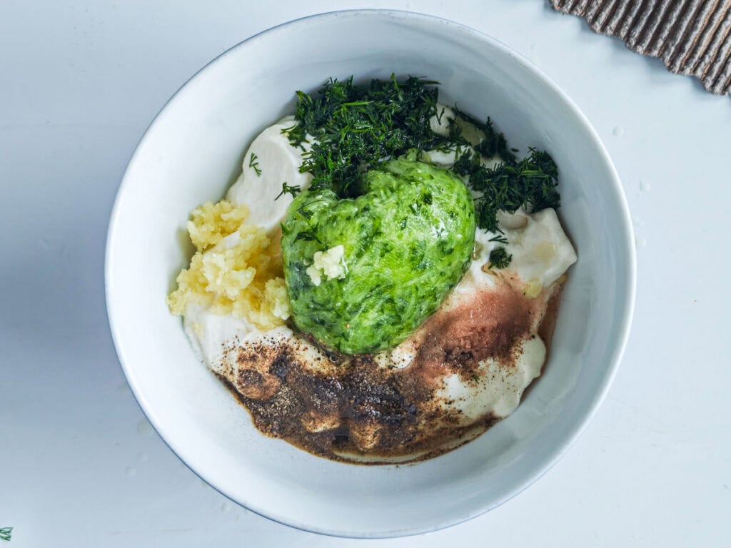 Grated cucumber, Greek yogurt, dill, garlic, lemon juice, oil, salt and pepper in a large white bowl.