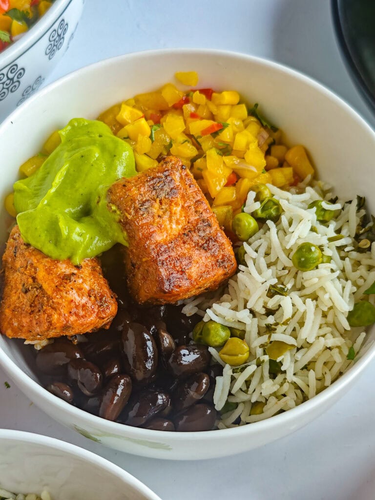 Salmon burrito bowl with taco-seasoned salmon, lime cilantro rice, mango corn salsa, black beans and avocado crema in a bowl.