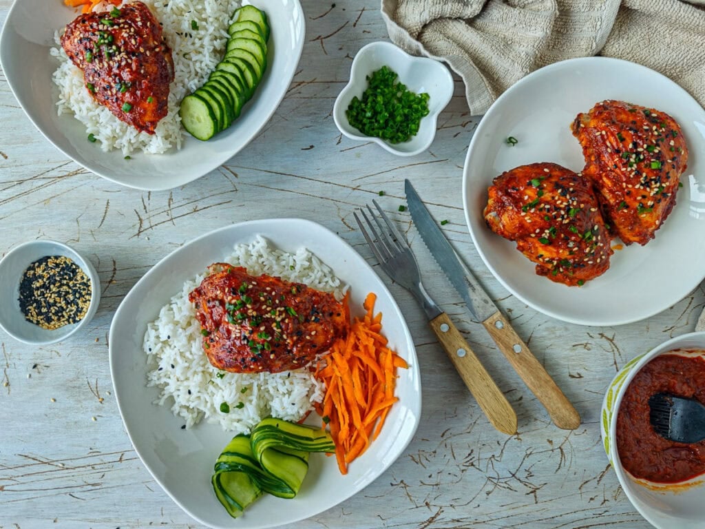 Easy Gochujang Chicken Thighs (Sticky & Oven-Baked) Gochujang chicken served with rice and vegetables on 2 different plates with sticky sauce, sesame seeds and spring onions by the side.