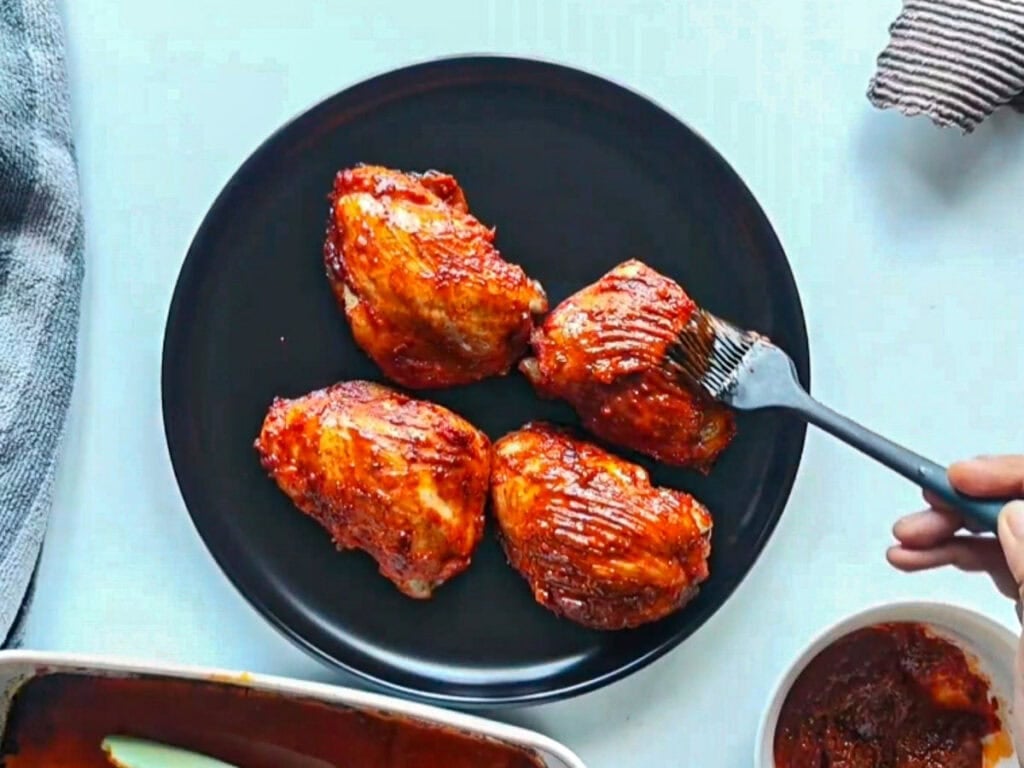 Sticky gochujang sauce being brushed on chicken thighs on a black plate.