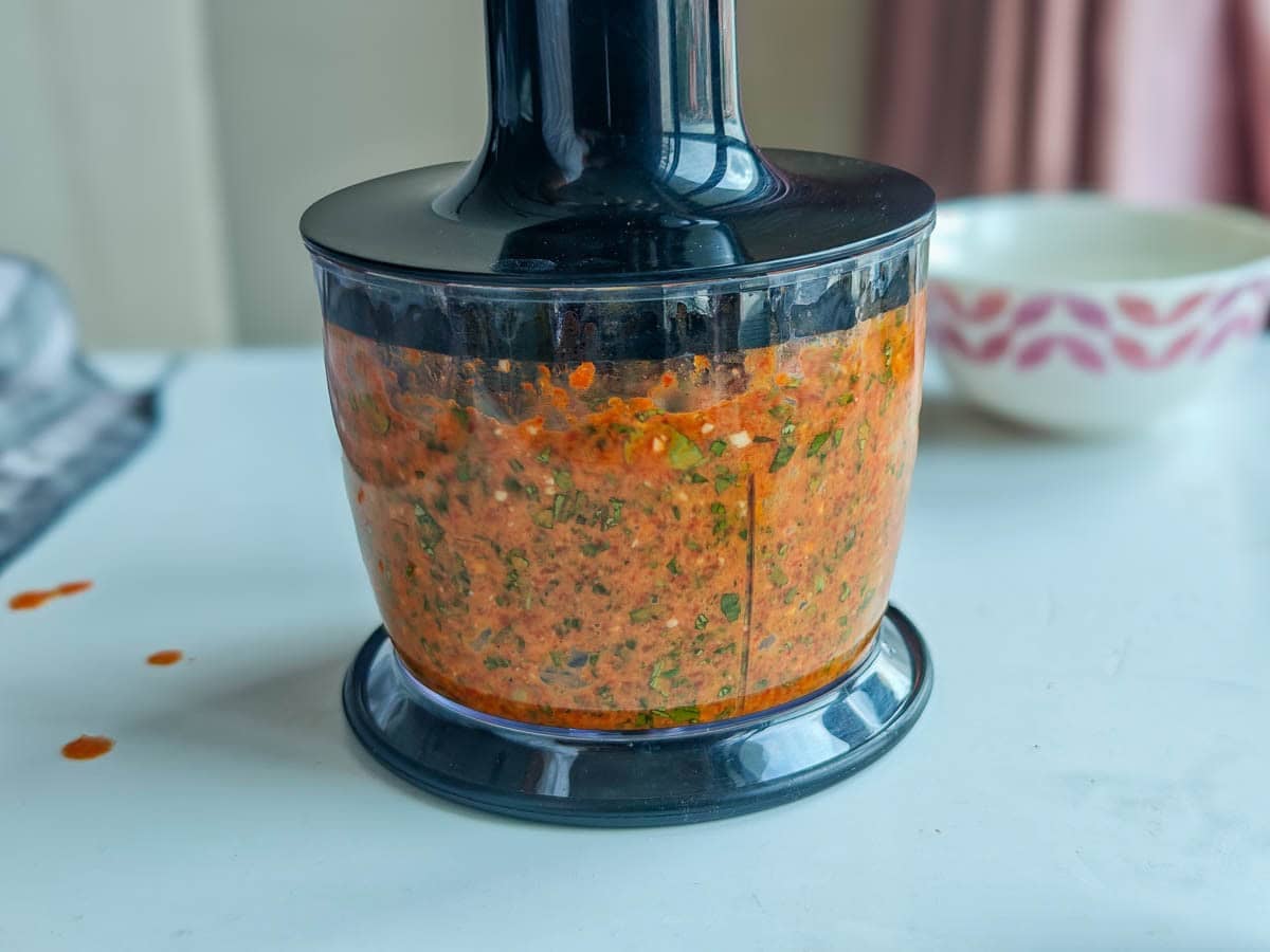 Easy Red Chimichurri Sauce with Roasted Peppers Herbs being blended with roasted red peppers and spices in an immersion blender.